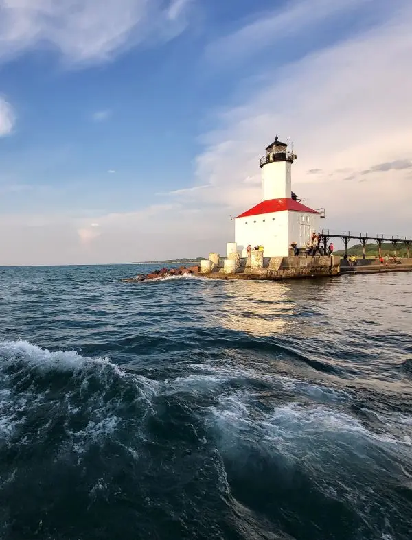 A charming lighthouse perched on a pier extends into the tranquil waters of the Great Lakes, captured during a serene spring evening. Visitors nearby enhance the calm yet adventurous spirit of a Midwest spring break destination by the water.