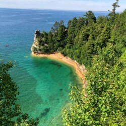 Cliffside view of a serene cove with crystal-clear turquoise waters along the coast of Lake Superior, framed by lush greenery. Kayakers enjoy the calm waters near a notable rock formation, making it an idyllic Midwest spring break destination for nature lovers and outdoor enthusiasts.