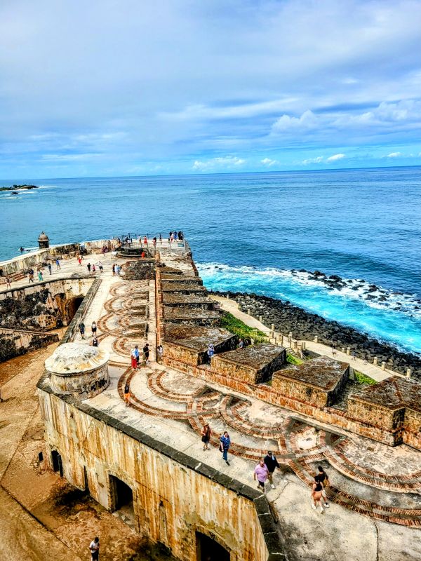 Aerial view of visitors exploring the historic fortification walls of San Juan, showcasing a labyrinth-like pattern on the ground, with the vast blue ocean extending to the horizon. This spot is a photographer's delight for capturing San Juan's rich history and oceanic backdrop.