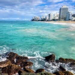 The turquoise waters of the Caribbean Sea wash over rocks at Condado Beach in Puerto Rico, with the skyline of San Juan in the background. This coastal scene is amazing for a Puerto Rico girls trip.