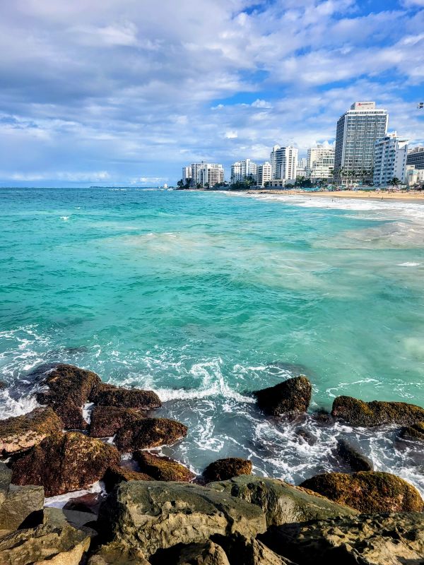 The turquoise waters of the Caribbean Sea wash over rocks at Condado Beach in Puerto Rico, with the skyline of San Juan in the background. This coastal scene is amazing for a Puerto Rico girls trip.