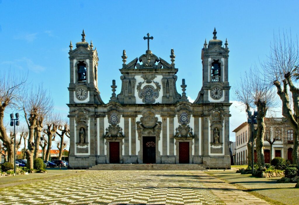 Baroque church of Bom Jesus de Matosinhos, a notable landmark on the route from Porto to Matosinhos, Portugal. Featuring twin bell towers and intricate façade details, it stands under a clear blue sky, fronted by a patterned cobblestone square and bare trees.