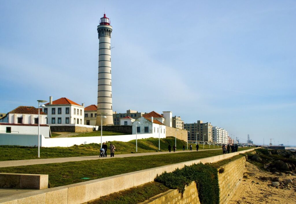 Iconic lighthouse in Matosinhos on the coastal journey from Porto, overlooking a beachside promenade. People are strolling along the walkway on a sunny day, with the backdrop of a clear blue sky and urban landscape.