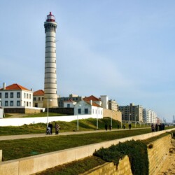 Iconic lighthouse in Matosinhos on the coastal journey from Porto, overlooking a beachside promenade. People are strolling along the walkway on a sunny day, with the backdrop of a clear blue sky and urban landscape.