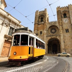 Historic Lisbon Cathedral with its twin Gothic towers, captured alongside the famous yellow Tram 28, a symbol of Lisbon's charming public transport, against a cobblestone street backdrop on a sunny day.