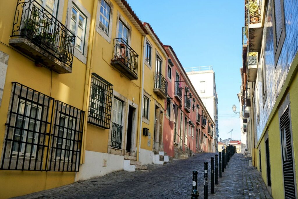 Quiet, sunlit alleyway lined with mustard and rose-colored buildings in Lisbon, reflecting the serene beauty ideal for a leisurely stroll on a Lisbon itinerary.