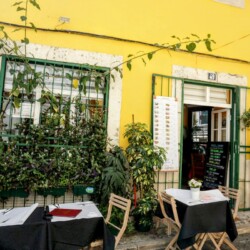 A cozy outdoor dining setup against a bright yellow wall of a quaint Lisbon restaurant, complete with lush greenery and a welcoming open door, a perfect meal stop for a Lisbon itinerary.