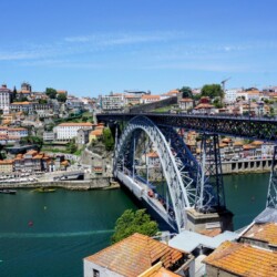 A captivating view of the iconic Dom Luís I Bridge arcing over the Douro River, with the historic Porto skyline rising in the background, under a clear blue sky.