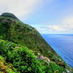 Lush greenery blankets the steep slopes of Madeira, with cultivated terraces and a small cluster of houses nestled near the coastline. The expansive blue Atlantic Ocean extends to the horizon, enhancing the scenic charm of a Madeira itinerary.