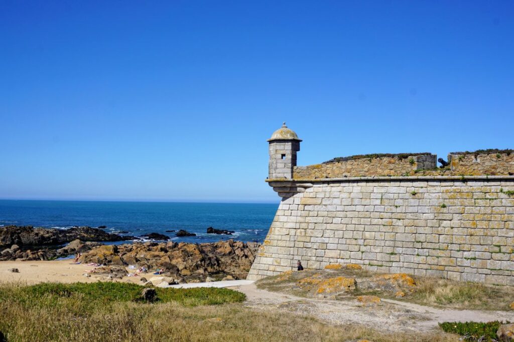 Historical fortification with a watchtower overlooking the rocky shores of Matosinhos near Porto. The serene Atlantic Ocean provides a beautiful contrast to the sunlit ancient stone walls, with visitors enjoying the beach below on a clear day.