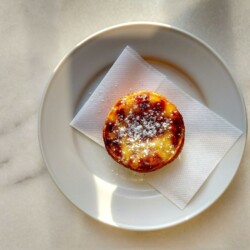 A single Pastel de Nata dusted with powdered sugar sits on a white plate, highlighted by natural sunlight.