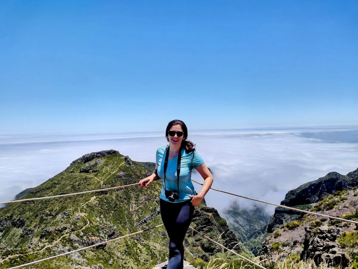 A smiling hiker posing at Pico do Arieiro, Madeira, with a breathtaking backdrop of mountain peaks rising above clouds. This image showcases a popular spot for those following a Madeira itinerary, emphasizing the island's dramatic scenery.