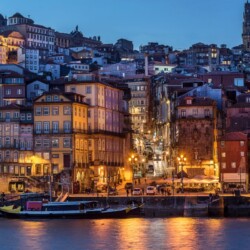 Twilight view of Porto's Ribeira district with layered, colorful houses ascending the hillside, lively street lights beginning to glow, and traditional Rabelo boats docked along the Douro River.