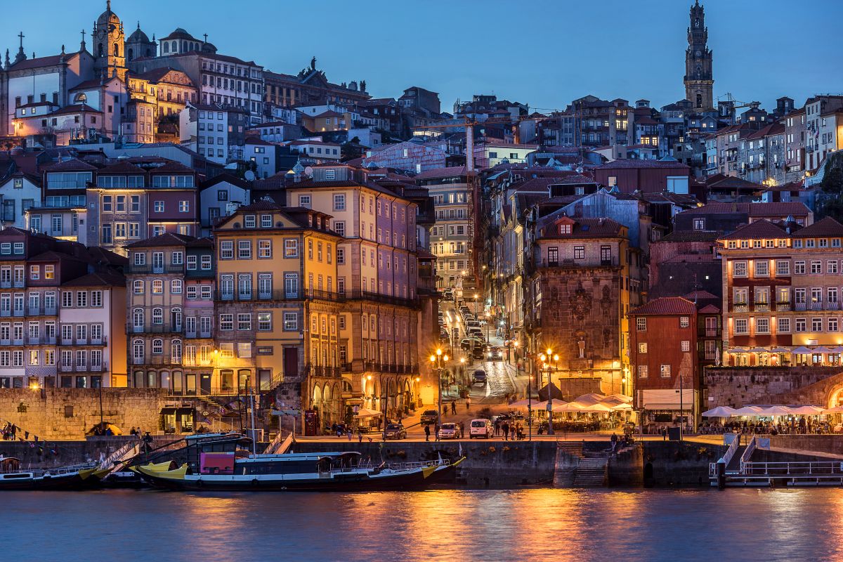 Twilight view of Porto's Ribeira district with layered, colorful houses ascending the hillside, lively street lights beginning to glow, and traditional Rabelo boats docked along the Douro River.