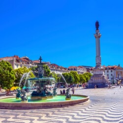 Rossio Square in Lisbon under the bright sun, showcasing the wavy-patterned cobblestones, central fountain, and the towering Column of Pedro IV, with people enjoying the lively urban space.