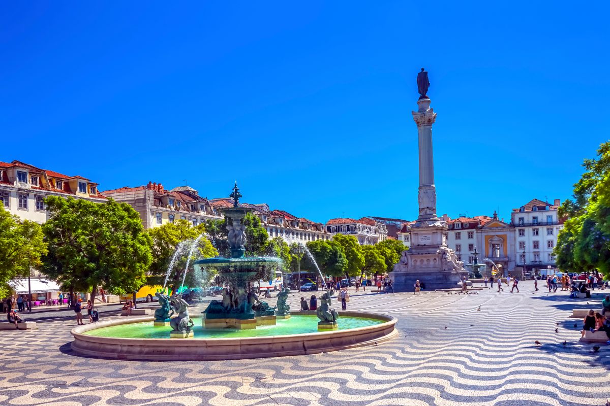 Rossio Square in Lisbon under the bright sun, showcasing the wavy-patterned cobblestones, central fountain, and the towering Column of Pedro IV, with people enjoying the lively urban space.