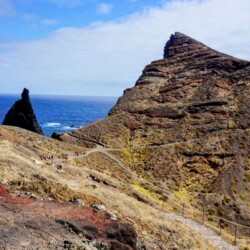 Hikers traverse a scenic trail at São Lourenço, Madeira, with a striking sea stack rising from the ocean and rugged cliffs forming a dramatic backdrop. The varied terrain, from red soil to green shrubs, illustrates the diverse natural beauty encountered on a Madeira itinerary.