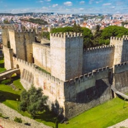 Aerial view of São Jorge Castle, a medieval fortress atop a hill in Lisbon, surrounded by lush greenery and offering a panoramic view of the city's colorful architecture under a clear sky. It is an iconic stop on a Lisbon itinerary.