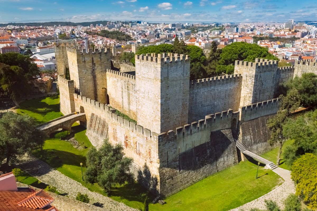 Aerial view of São Jorge Castle, a medieval fortress atop a hill in Lisbon, surrounded by lush greenery and offering a panoramic view of the city's colorful architecture under a clear sky. It is an iconic stop on a Lisbon itinerary.