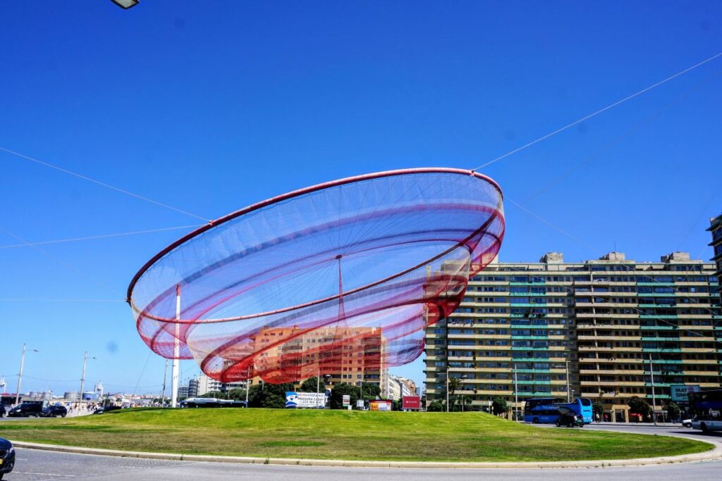 The dynamic 'She Changes' sculpture in Matosinhos, a striking landmark on the coastal route from Porto. This large, red, net-like structure contrasts against the deep blue sky, adding a modern touch to the urban landscape.