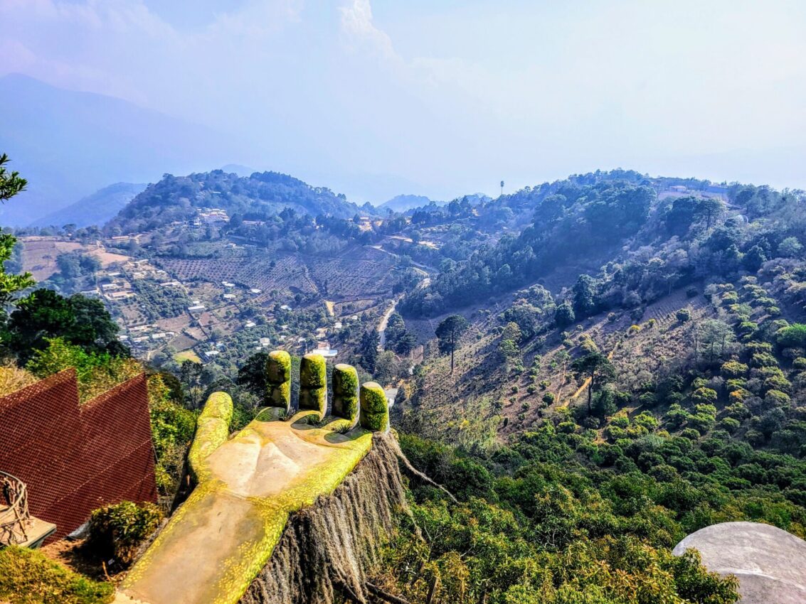 Sweeping view of the verdant valleys near Hobbitenango, Guatemala, with a whimsical oversized hand sculpture protruding from a cliff, epitomizing the unique attractions to discover on a Hobbitenango day trip.