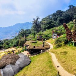 Panoramic perspective of Hobbitenango's terraced landscape with thatched structures and vibrant flower installations, inviting visitors to enjoy the scenic beauty on a day trip.