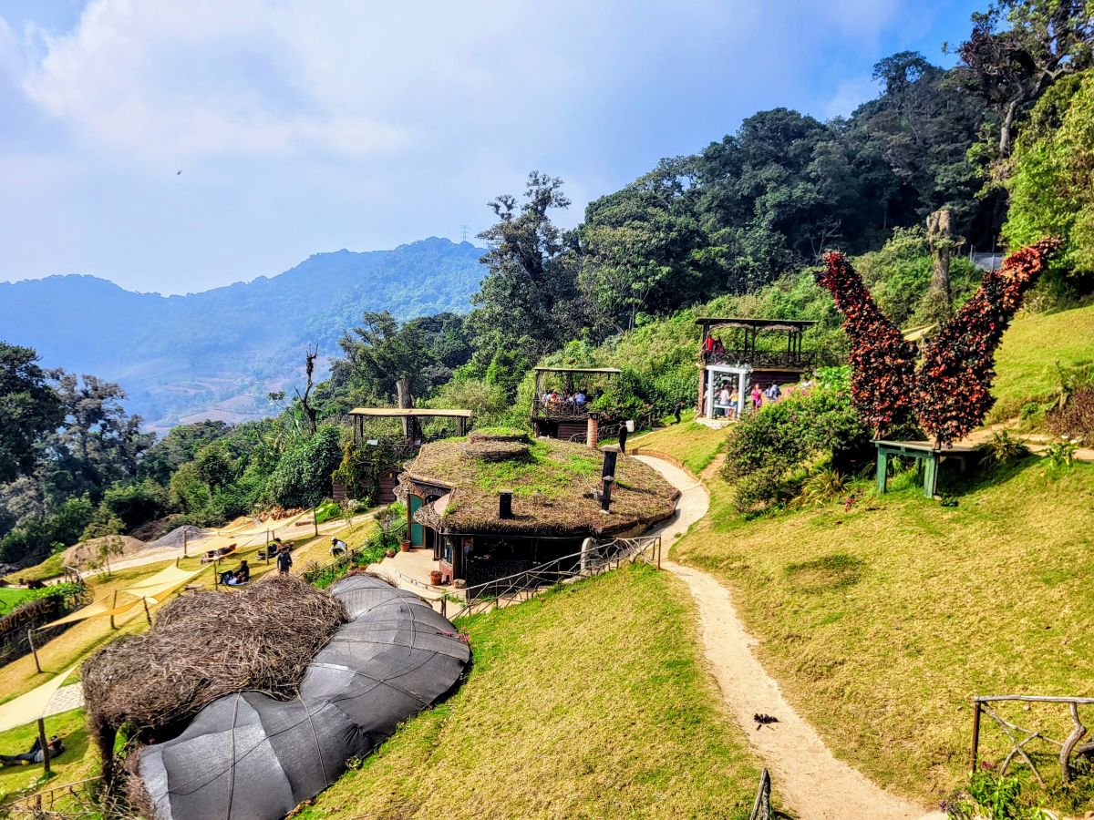Panoramic perspective of Hobbitenango's terraced landscape with thatched structures and vibrant flower installations, inviting visitors to enjoy the scenic beauty on a day trip.