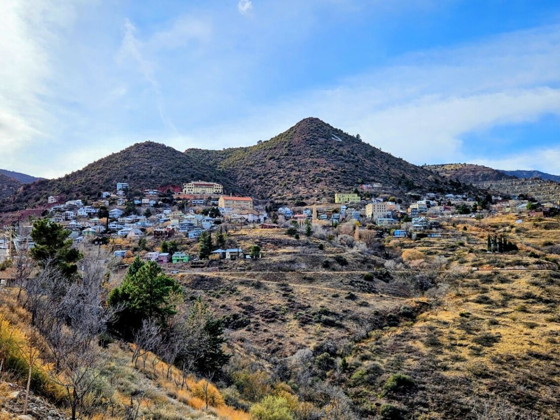 A hillside viewpoint in Jerome, Arizona. Colorful buildings dot the cliffside mountain, surrounded by a blue sky with clouds.