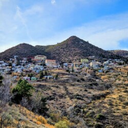 A hillside viewpoint in Jerome, Arizona. Colorful buildings dot the cliffside mountain, surrounded by a blue sky with clouds.