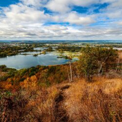 Breathtaking autumn vista from a high vantage point on a Midwest road trip, overlooking a patchwork of fall foliage, tranquil water bodies, and distant towns.