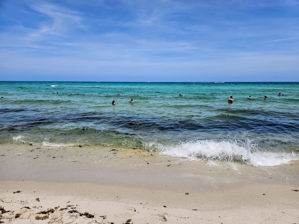 Blue waves crash against the sand along the Miami Beach shoreline, surrounded by blue skies on a clear day.
