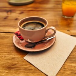 A pink, ceramic coffee mug on a small plate with a small spoon at a cafe in Porto. There is a glass of orange juice to the side. The table is wooden and rustic.