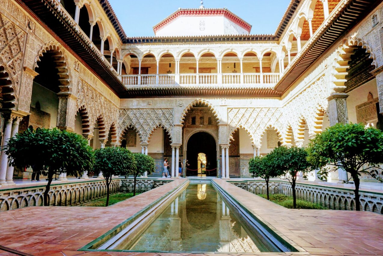 Courtyard of the Maidens in the Royal Alcázar with ornate Moorish arches, carved wooden balconies, and a long reflective pool, a serene stop on a Seville tour.