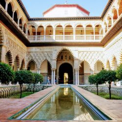 Courtyard of the Maidens in the Royal Alcázar with ornate Moorish arches, carved wooden balconies, and a long reflective pool, a serene stop on a Seville tour.