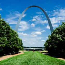 Iconic Gateway Arch on a sunny day framed by lush green trees, with a clear blue sky overhead, a perfect stop on a Midwest road trip.